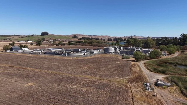 Drone pull out shot of the Petaluma Water Recycling Facility in California, showing the plant's circular clarifiers and treatment tanks set against a dry, rural landscape with rolling hills