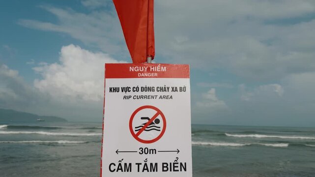 Red flag flutters over beach safety sign warning of rip currents and prohibited swimming. Rolling surf and distant islands form coastal backdrop to this Vietnamese public safety notice. Danang