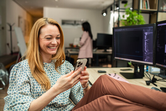 Smiling woman using smartphone with feet up at office desk