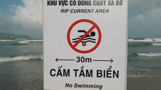Rip current warning sign stands beside rough beach water, prohibiting swimming within 30 meters. Bilingual safety notice marks dangerous surf as waves break beneath cloudy coastal skies. Danang
