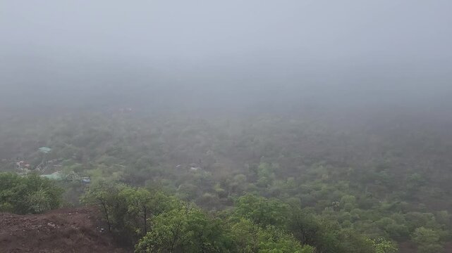 Dense white fog and heavy mist slowly drift over lush green forested mountain hills during overcast rainy weather, creating a dramatic and moody atmospheric landscape b-roll shot.