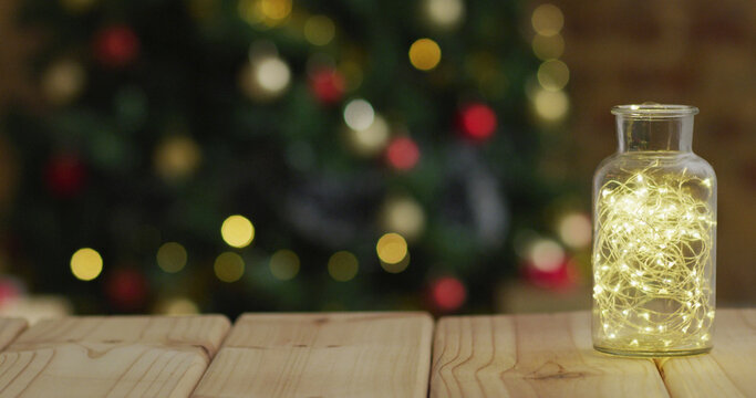 Image of wooden table with fairy lights in jar over defocussed christmas tree with baubles