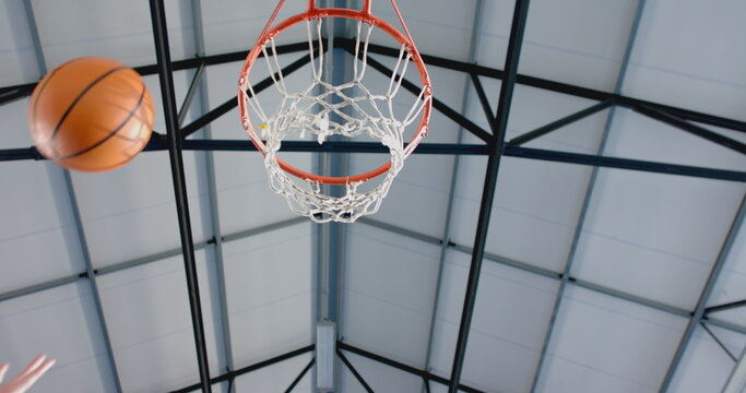 Flying basketball passing by hand toward hoop under steel truss in gym, net rippling