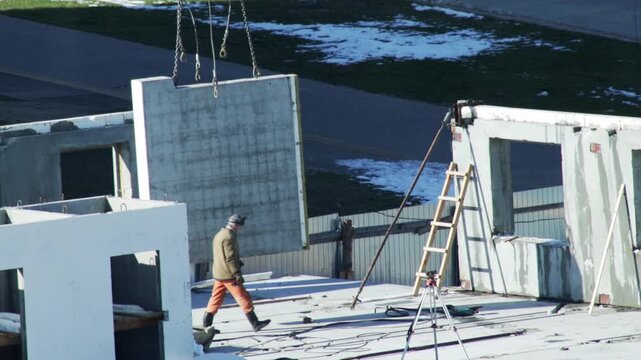 Construction of a residential building site, heavy concrete panel lifted by crane for wall assembly, worker preparing for installation, development progress on cold winter day with snow patches