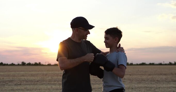 Father hugs son after boxing training at sunset. They touch gloved fists in open field, sharing respect, support, and pride. Family fitness and mentorship create warm outdoor sports moment.