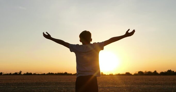 Boy stands with arms outstretched in field at sunset, enjoying freedom and summer evening. Silhouette shows joy, happiness and carefree childhood. Inspiring scene suits tourism, wellness and growth.