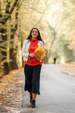 Smiling woman in red blouse and white jacket holding autumn leaves while walking along a wooded paved path in a sunlit fall forest portrait