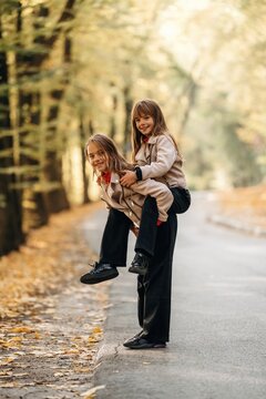 Two smiling girls giving a piggyback ride on a tree lined path with fallen leaves and soft sunlight, playful outdoor childhood scene in autumn woodland