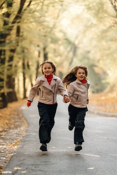 Two young girls running hand in hand along a tree lined park path in autumn, wearing matching jackets and scarves, outdoor childhood portrait