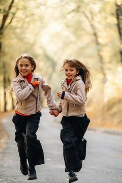 Two girls holding hands while running together on a tree lined paved path in autumn light, wearing casual jackets and dark pants outdoors