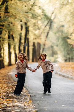 Two children holding hands while walking along a tree lined road covered with autumn leaves in a softly blurred outdoor portrait scene