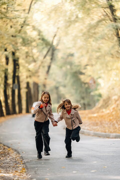 Two schoolgirls holding hands while running along a tree lined paved path in autumn, wearing casual jackets and backpacks in a sunlit outdoor scene