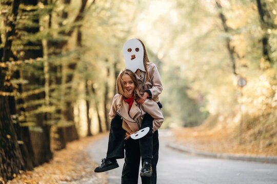 Parent carrying a child in a white costume mask while another child holds a painter palette on a sunlit forest road, autumn outdoor portrait