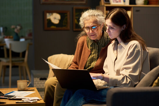 Young Caucasian woman helping senior Hispanic woman managing finances on laptop at home. Useful for digital literacy, elder care, online banking, and family assistance marketing