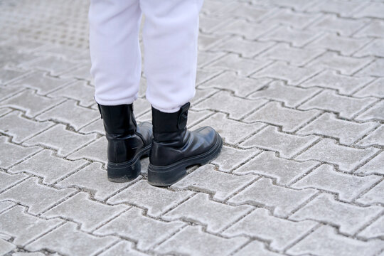 Female legs in black leather boots standing on dry paving tiles covered with white rock salt residue and grit for winter traction. Female feet in black boots on salt treated city sidewalk