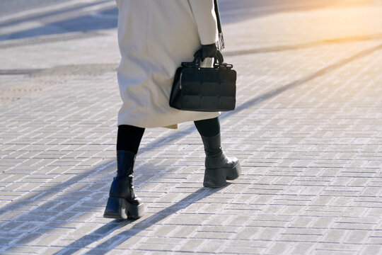 Female legs in white coat and black boots walking on street pavement covered in traction grit and salt. Woman in boots taking steps on dry sidewalk treated with rock salt for anti slip winter traction