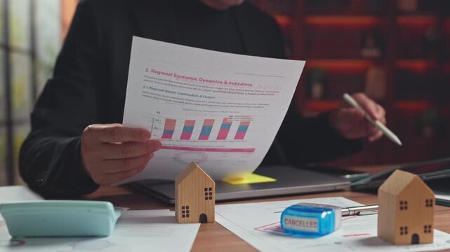 Businessman analyzes regional economic indicators report, wooden house model cancelled stamp in office. Real estate bankruptcy, mortgage foreclosure crisis, property investment audit.