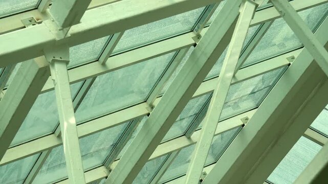 Glass ceiling framework showcases complex geometric steel beams supporting large roof windows. Natural daylight filters through translucent glass panels, highlighting industrial architecture.