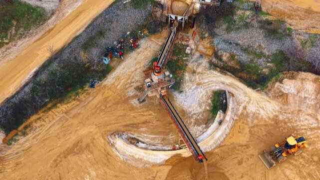 Top down view of sand mining conveyor system. Metal conveyor belt deposits sand while a yellow wheel loader stands on sandy ground.