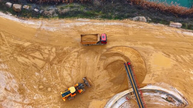 Top down view of dump truck and wheel loader. Red dump truck loaded with sand stands near a yellow loader and conveyor belt.