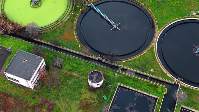 Aerial shot of water treatment plant featuring circular settling tanks. Top view depicts green algae basin alongside black sedimentation pools and administrative building.