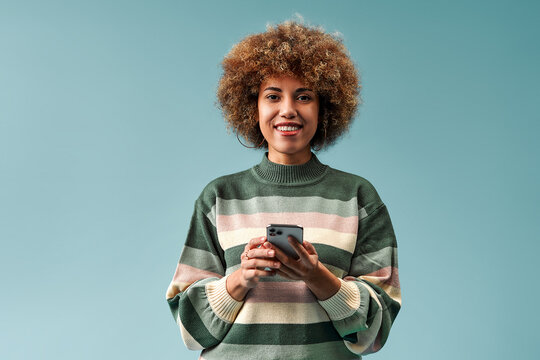 Smiling young woman with curly hair holding a smartphone while wearing a striped knit sweater against a clean teal background.