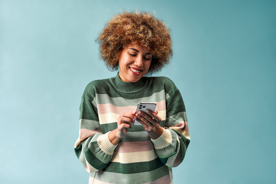 Smiling young woman with curly hair using a smartphone while wearing a striped sweater against a plain blue studio background.