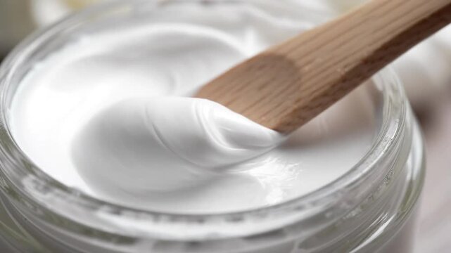 Close-up of a wooden spatula scooping luxurious white cream from a glass jar, highlighting smooth texture and natural beauty products.