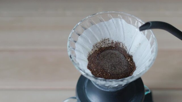 Drip coffee, close up of hot water pouring from gooseneck kettle into glass dripper with paper filter and ground coffee pour over brewing