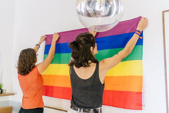 Two women hanging LGBTQ+ rainbow flag on wall