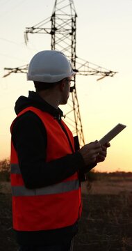 Electrical engineer inspects power transmission tower at sunset. Worker in safety helmet uses tablet for grid maintenance and field inspection. Reliable energy service supports utility operations.
