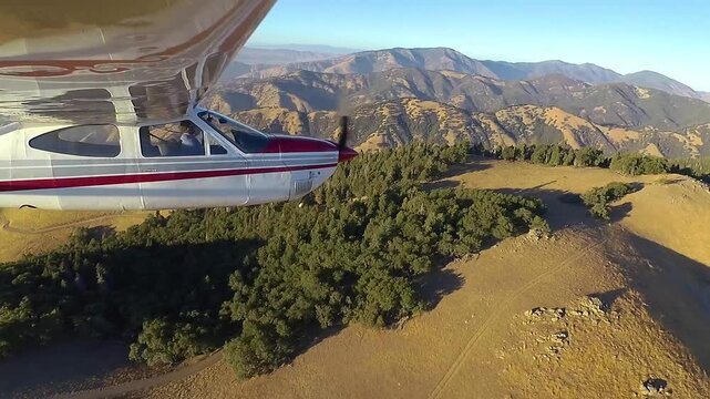 Profile view of a small airplane flying along and then making a steep bank over a high and partially forrested ridge in California's Tehachapi Mountains in the late afternoon.
