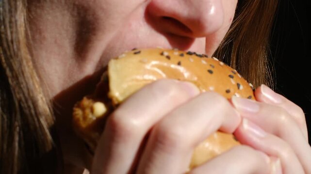 Hungry woman with red lipstick biting and chewing a juicy sesame chicken burger with sauce, savoring a tasty fast food meal in close up against a black background