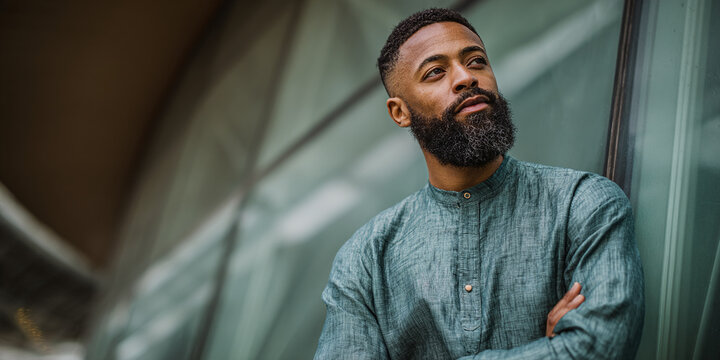Portrait of a bearded Black man looking upwards, thoughtful, with arms crossed in front of a modern building, signifying contemplation and urban life