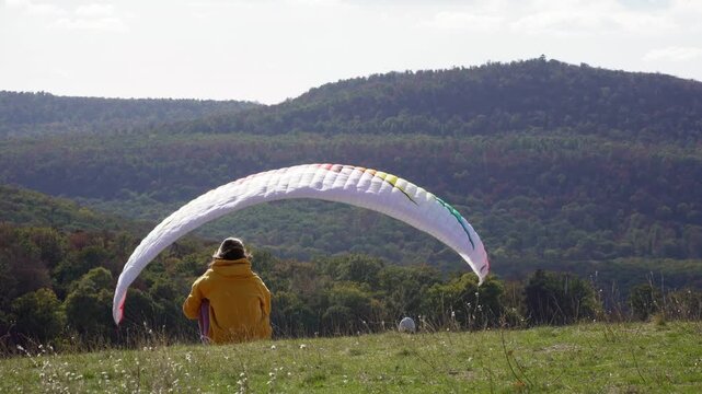Person sitting beside an inflated paraglider canopy on a grassy hill overlooking autumn forested mountains