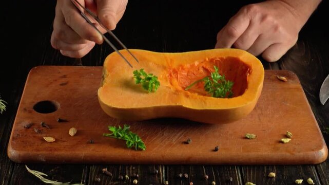 Hands arranging fresh green herbs inside a halved roasted butternut squash on a wooden cutting board with tweezers and scattered spices, overhead food preparation scene