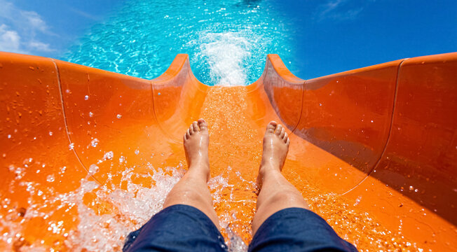 First person view of legs sliding down an orange water slide into a blue swimming pool, splashing water. Summer vacation, summer fun, water park activity.