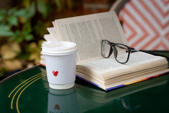Cozy coffee break on a glass table with a book, glasses, and a takeaway coffee cup decorated with a red heart on an outdoor cafe terrace