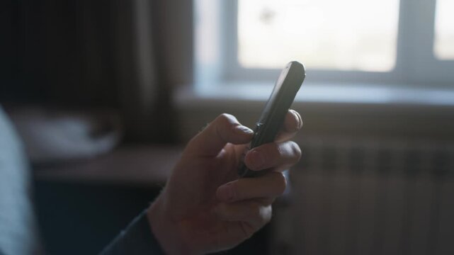 Person presses buttons on a vintage cellphone indoors in a focused close up view. Soft natural daylight illuminates the hands near a window with gentle selective focus.