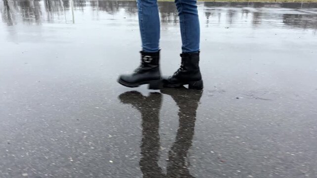 Female feet in boots going on wet asphalt road at spring or autumn. Legs of young woman stepping on puddles at rainy day. Concept of tranquility scene. Slow motion
