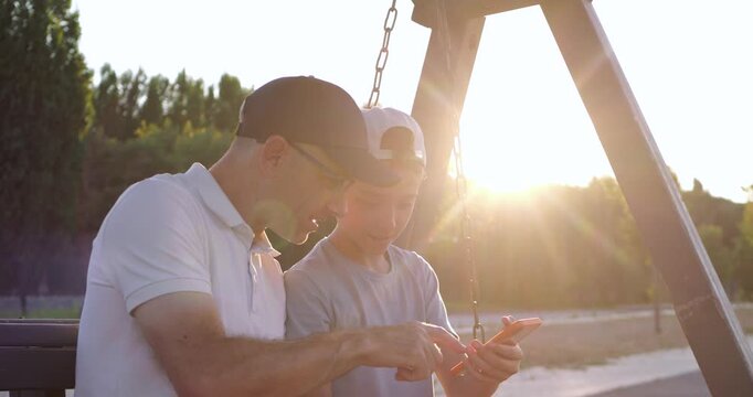 Father and son share smartphone on playground swing. Dad and child discuss technology during warm evening sunset. Family bonding and digital learning create authentic parenting lifestyle footage.