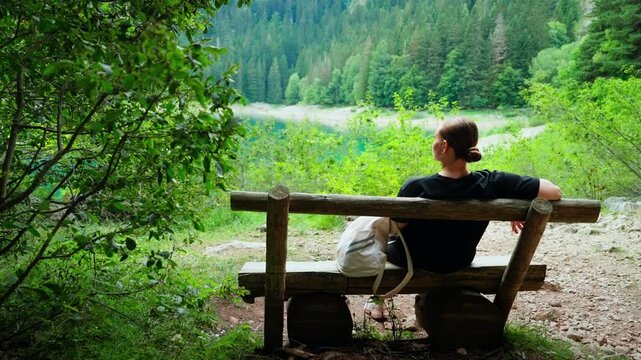 Woman resting on a rustic wooden bench quietly admiring a tranquil forest landscape with distant mountain peaks and serene lake in a peaceful nature retreat