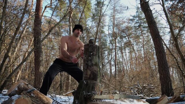 Shirtless young man chopping firewood in a sunny winter forest. Muscular guy working with axe in snowy woodland. Concept of strength, masculine, energy and harmony with nature during cold season