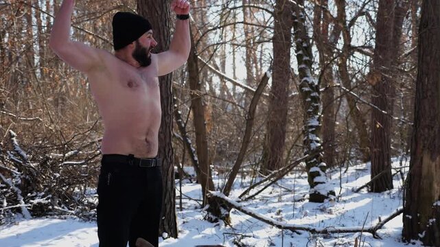 Shirtless muscular man plants an axe in stump at snowy forest. Strong guy flexes his biceps in playful stance showing endurance, power and confidence. The scene blends outdoor activity and strength