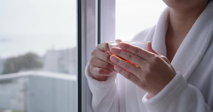 Woman is standing by window adjusting grip and bringing glass teacup to lips, then sipping tea