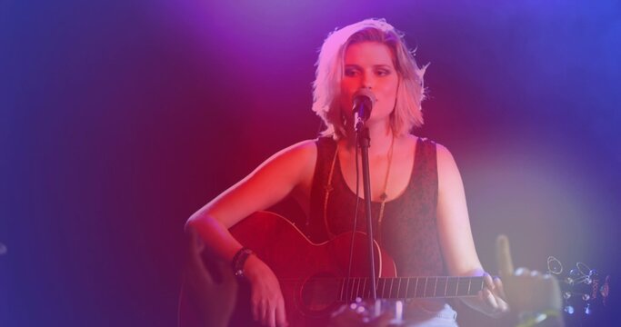Singing teen musician playing on club stage, with acoustic guitar and mic, wearing dark tank top