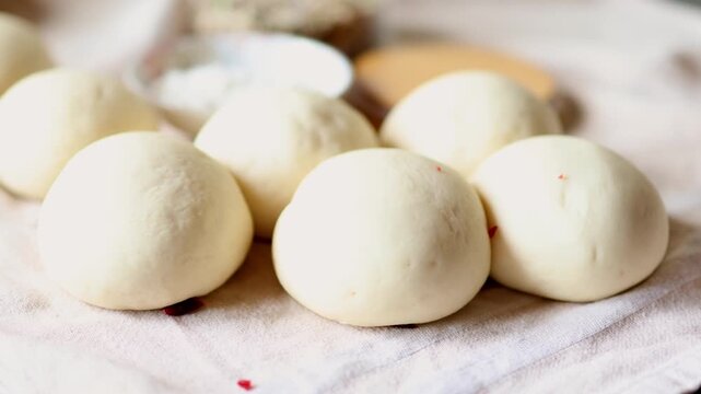 Timelapse of round yeast dough balls rising on a linen towel beside a bowl of flour. Concept of proofing dough, patient baking and the magic of fermentation in homemade bread making at home.