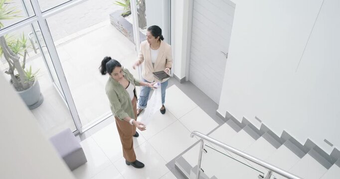 Asian women partners entering glass door, unrolling plans, checking tablet, pointing to align foyer