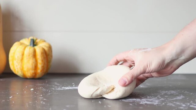 Hands kneading yeast dough on a floured surface with a yellow pumpkin and a squash in the background, 4k real time footage. Concept of traditional bread making and cozy autumn home cooking.