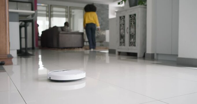 African American couple woman in yellow walking past partner sitting starting robot vacuum on tiles